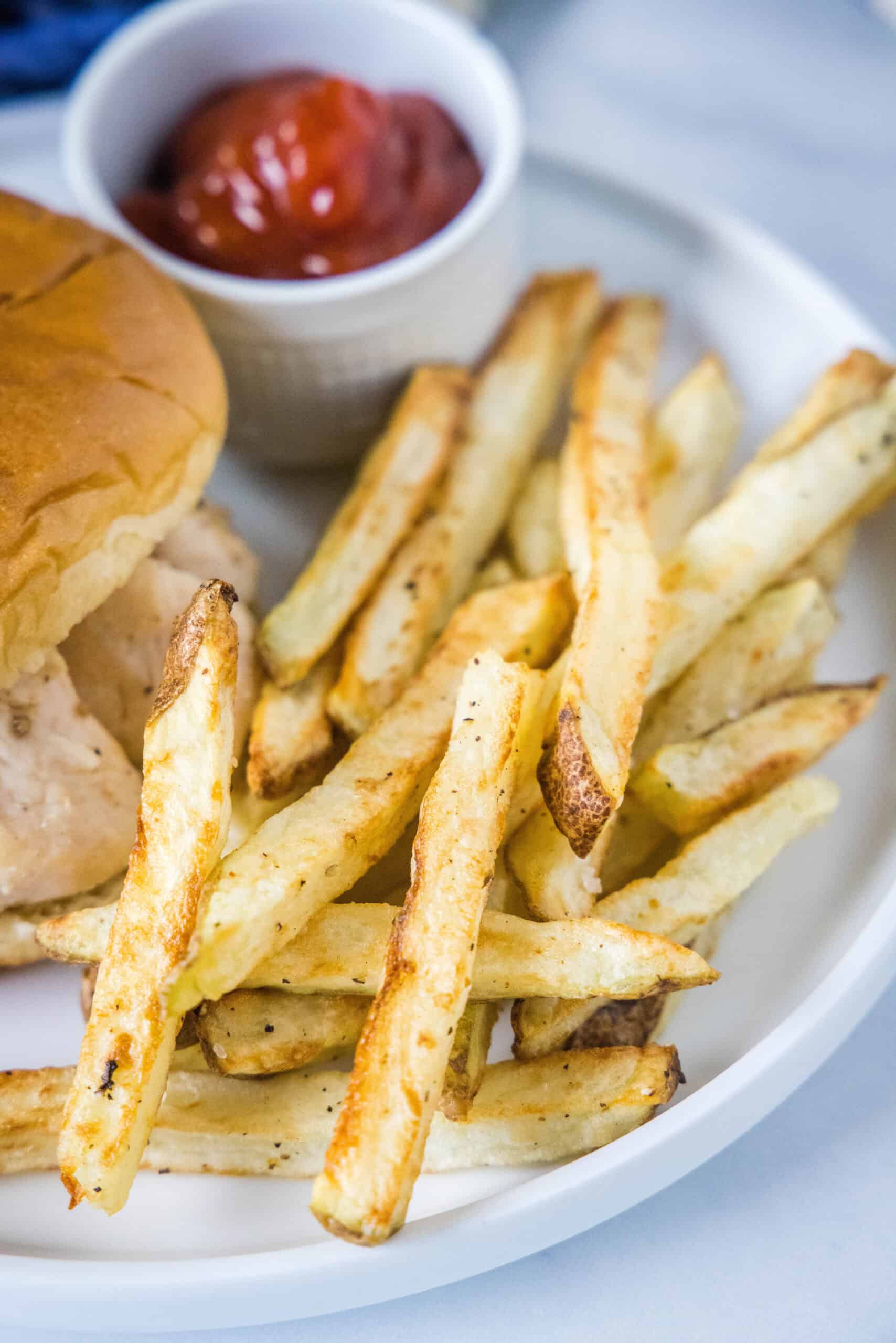 Homemade air fryer French fries are just as crispy as regular fries! Make them in a fraction of the time, with any seasonings you’d like. Homemade air fryer French fries next to a burger and a small ramekin filled with ketchup on a plate.