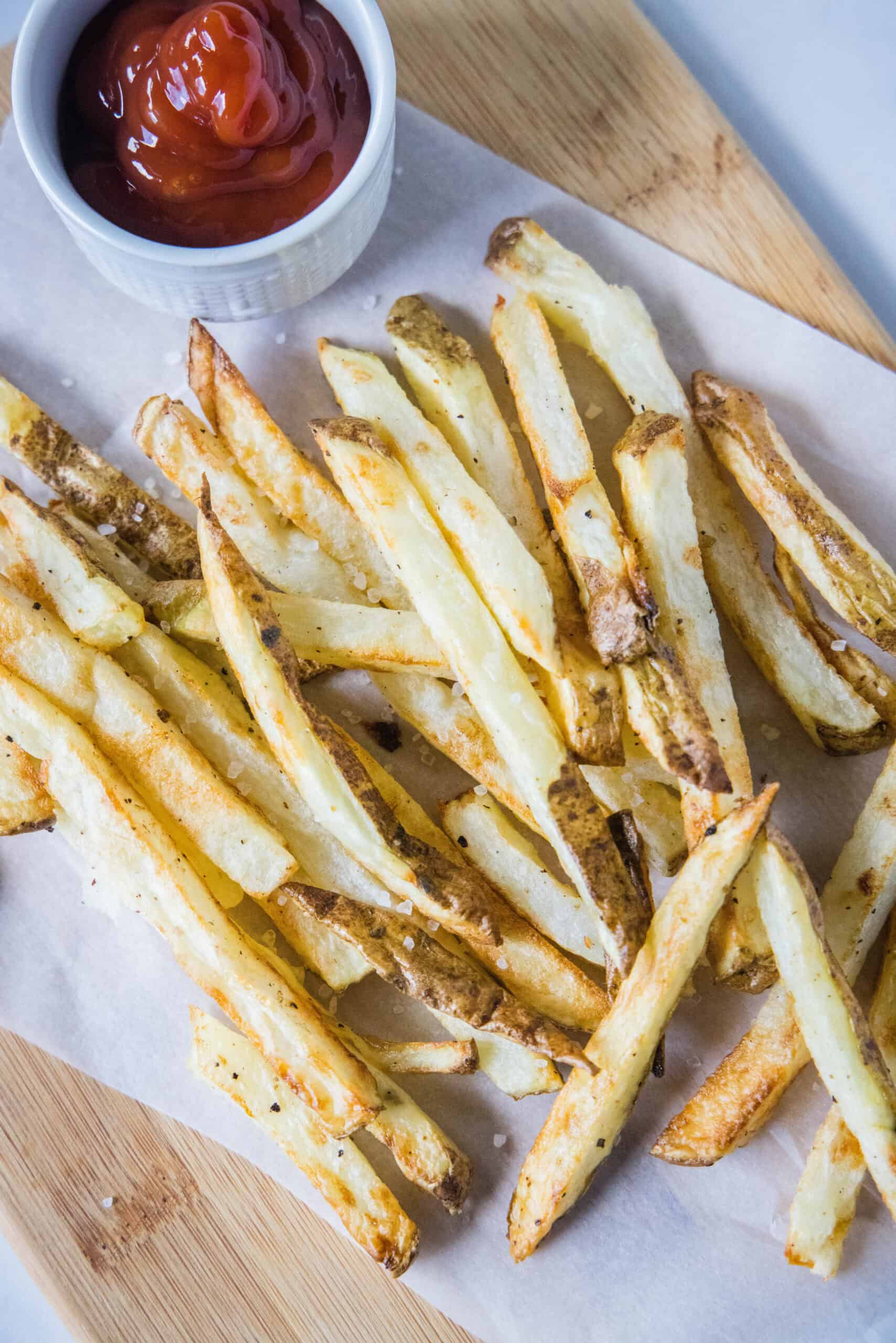 Homemade air fryer French fries are just as crispy as regular fries! Make them in a fraction of the time, with any seasonings you’d like. Overhead view of air fryer French fries on a sheet of parchment paper over a wooden cutting board, next to a small ramekin of ketchup.