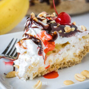 A slice of banana split pie drizzled with chocolate sauce and strawberry sauce, topped with chopped peanuts and cherries on a plate.