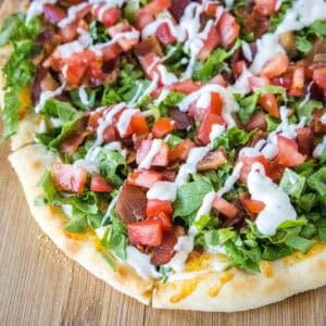 Close up of a BLT pizza topped with a drizzle of aioli on a wooden cutting board.