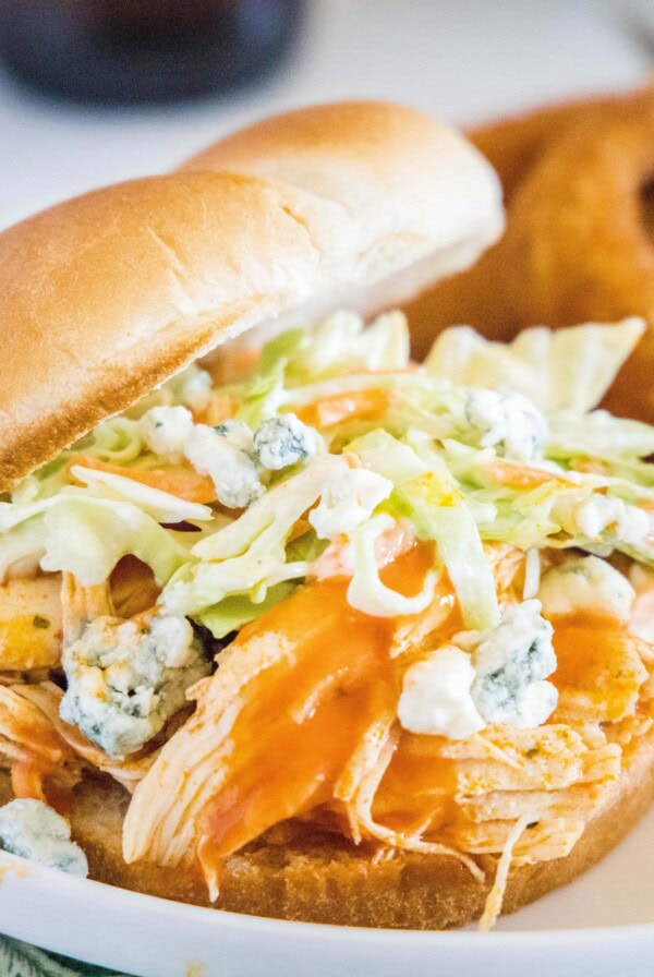 A buffalo chicken sandwich on a plate, topped with coleslaw and blue cheese, with onion rings in the background