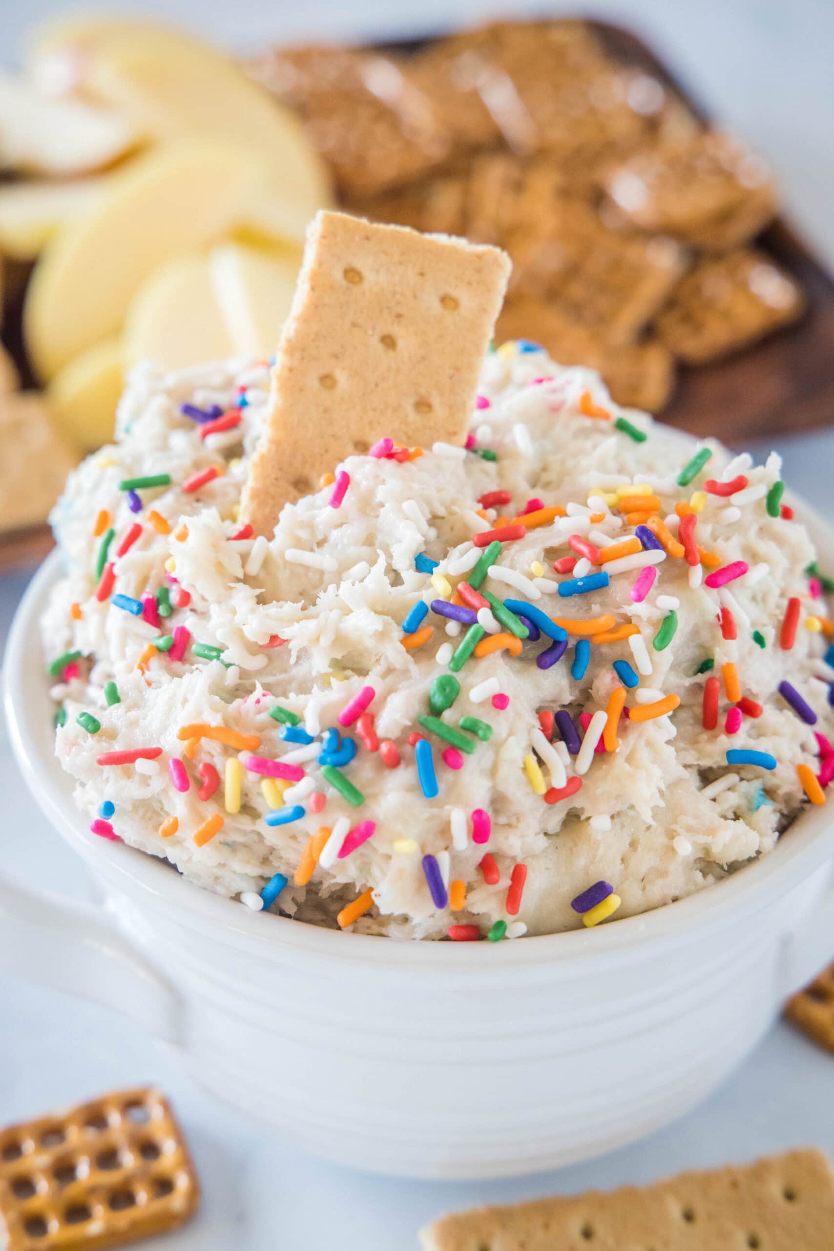 Fluffy cake batter dip is an easy no-bake dessert made from cake mix and brimming with sprinkles. It tastes just like funfetti cake batter! Cake batter dip in a white bowl with a cookie stuck into the top, and a platter of cookies in the background.