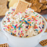 Cake batter dip in a white bowl with a cookie stuck into the top, and a platter of cookies in the background.
