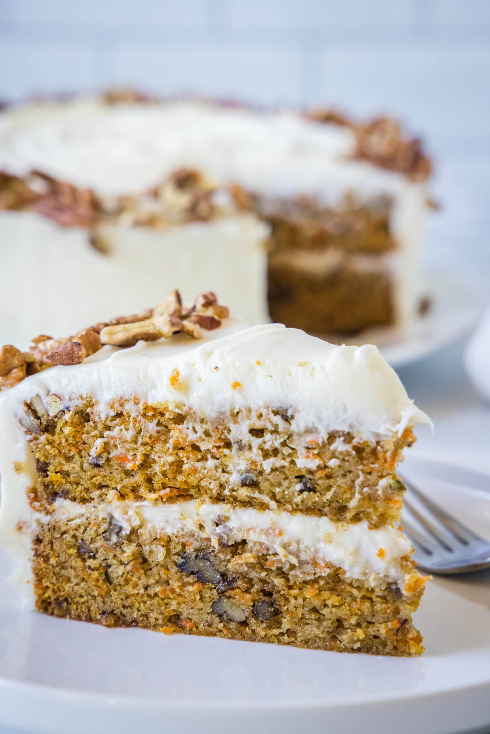 The best homemade carrot cake! Made from moist cake layers loaded with crunchy pecans, filled and frosted with sweet cream cheese frosting. A slice of frosted carrot cake on a white plate next to a fork, with the rest of the cake in the background.
