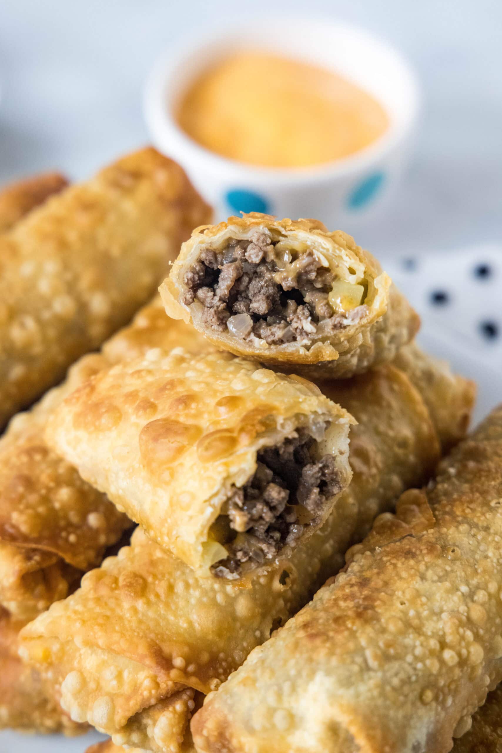 Looking for a fun and easy appetizer recipe? Try these cheeseburger egg rolls complete with creamy, tangy homemade burger sauce for dipping! Close up of cheeseburger egg rolls stacked on a platter, with one egg roll broken in two and a bowl of dipping sauce in the background.