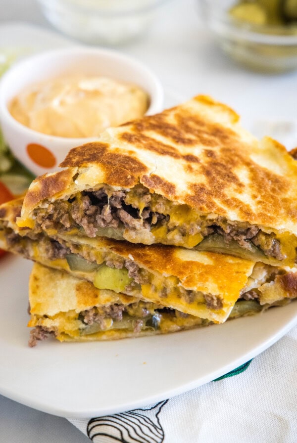 Three slices of cheeseburger quesadilla stacked on a plate, next to a bowl of burger sauce.