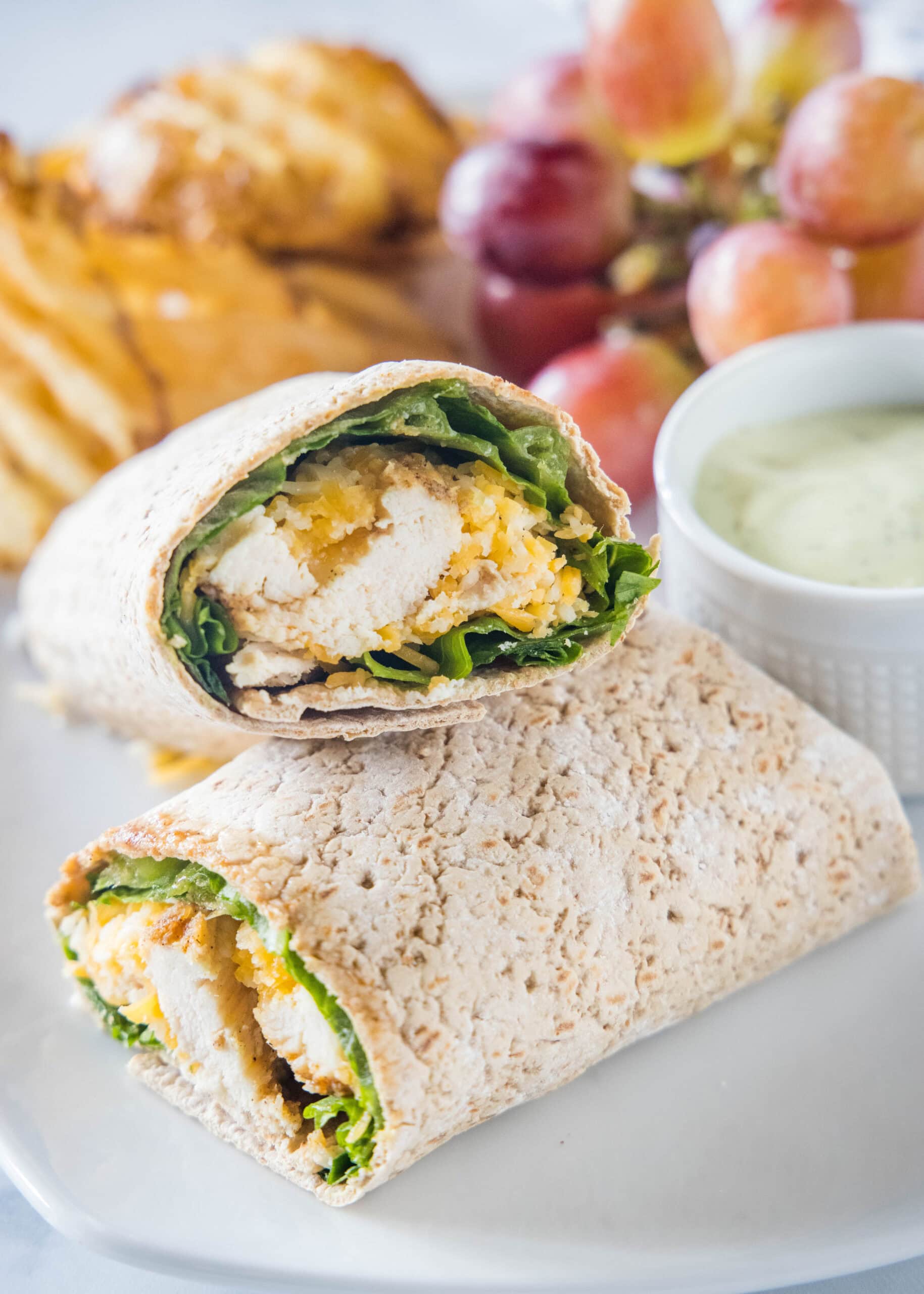 This easy Chick Fil A Cool Wrap recipe is the best excuse to skip the drive-thru! Enjoy this copycat at home with your favorite dipping sauce. Close up of two halves of a Chick Fil A Cool Wrap on a plate next to a bowl of dipping sauce, grapes, and potato chips.