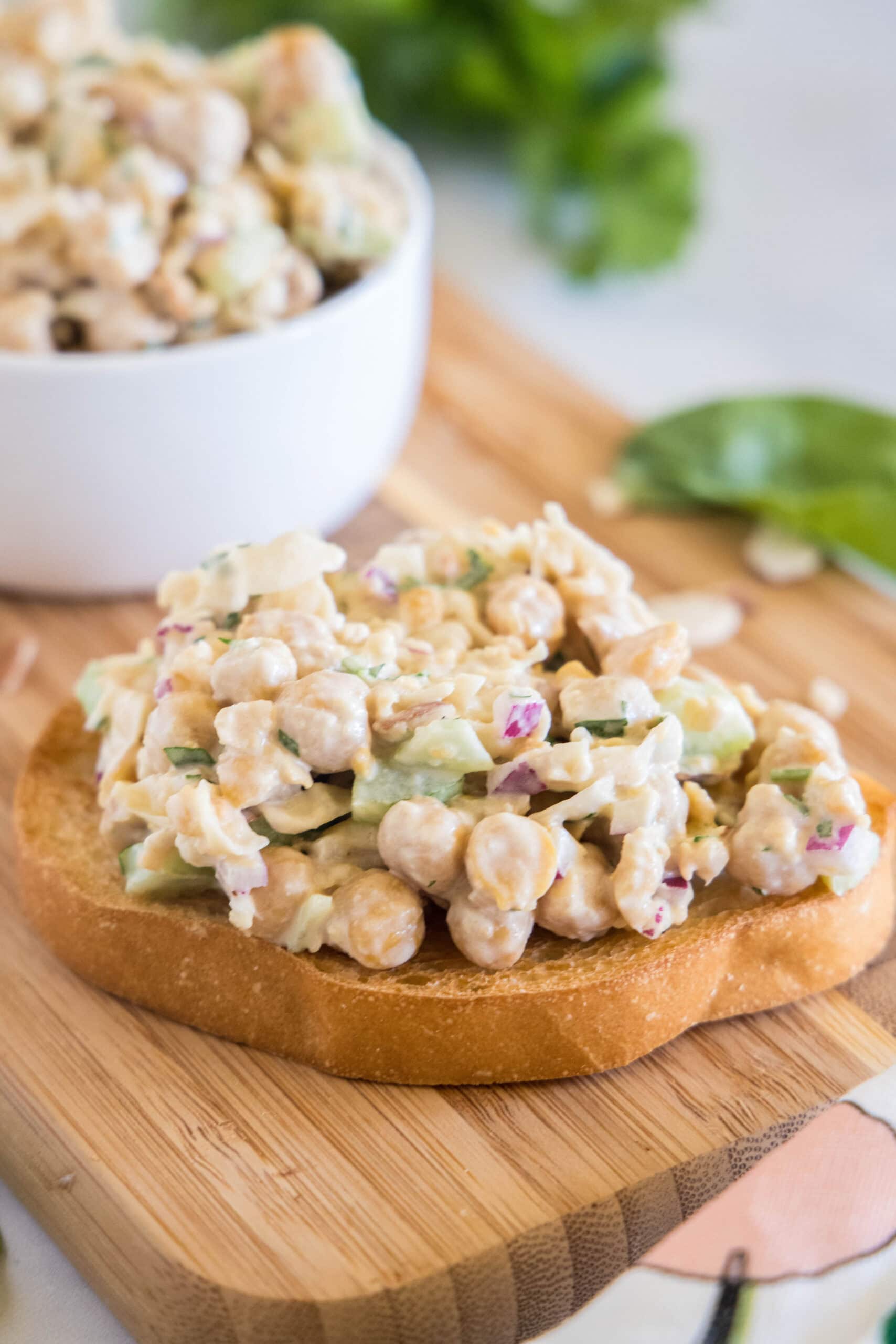 Chickpea chicken salad is a delicious and high-protein chickpea recipe that’s fresh, quick to make, and perfect for vegetarian lunches! Chickpea chicken salad served on a slice of bread on a wooden cutting board, with a bowl of chickpea salad in the background.
