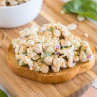 Chickpea chicken salad served on a slice of bread on a wooden cutting board, with a bowl of chickpea salad in the background.