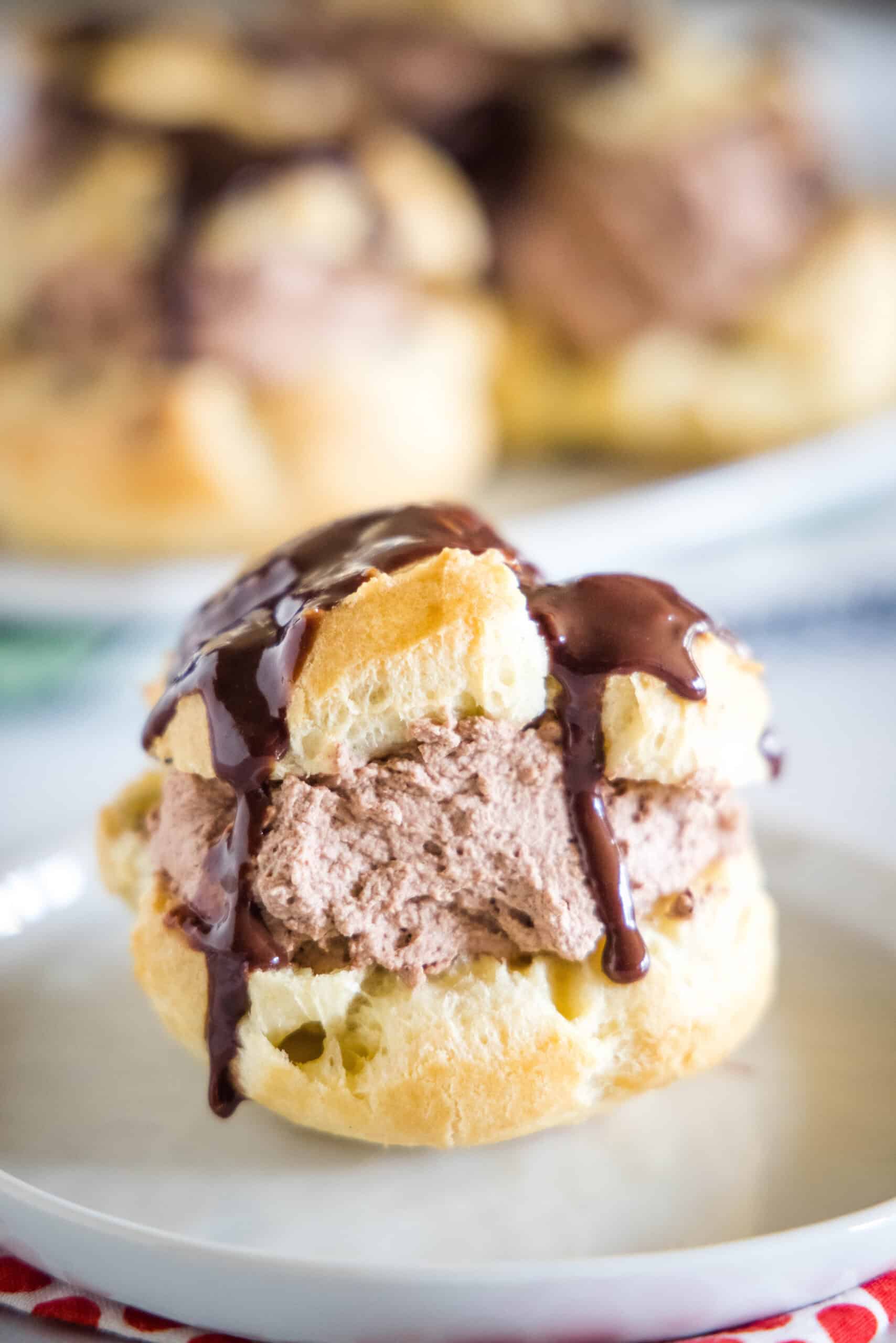 These chocolate cream puffs (profiteroles) are a chocolate lover’s dream, filled with fluffy chocolate whipped cream and topped with ganache. A chocolate cream puff topped with chocolate ganache on a white plate, with more cream puffs on a plate in the background.