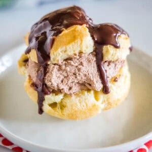 A chocolate cream puff topped with chocolate ganache on a white plate.