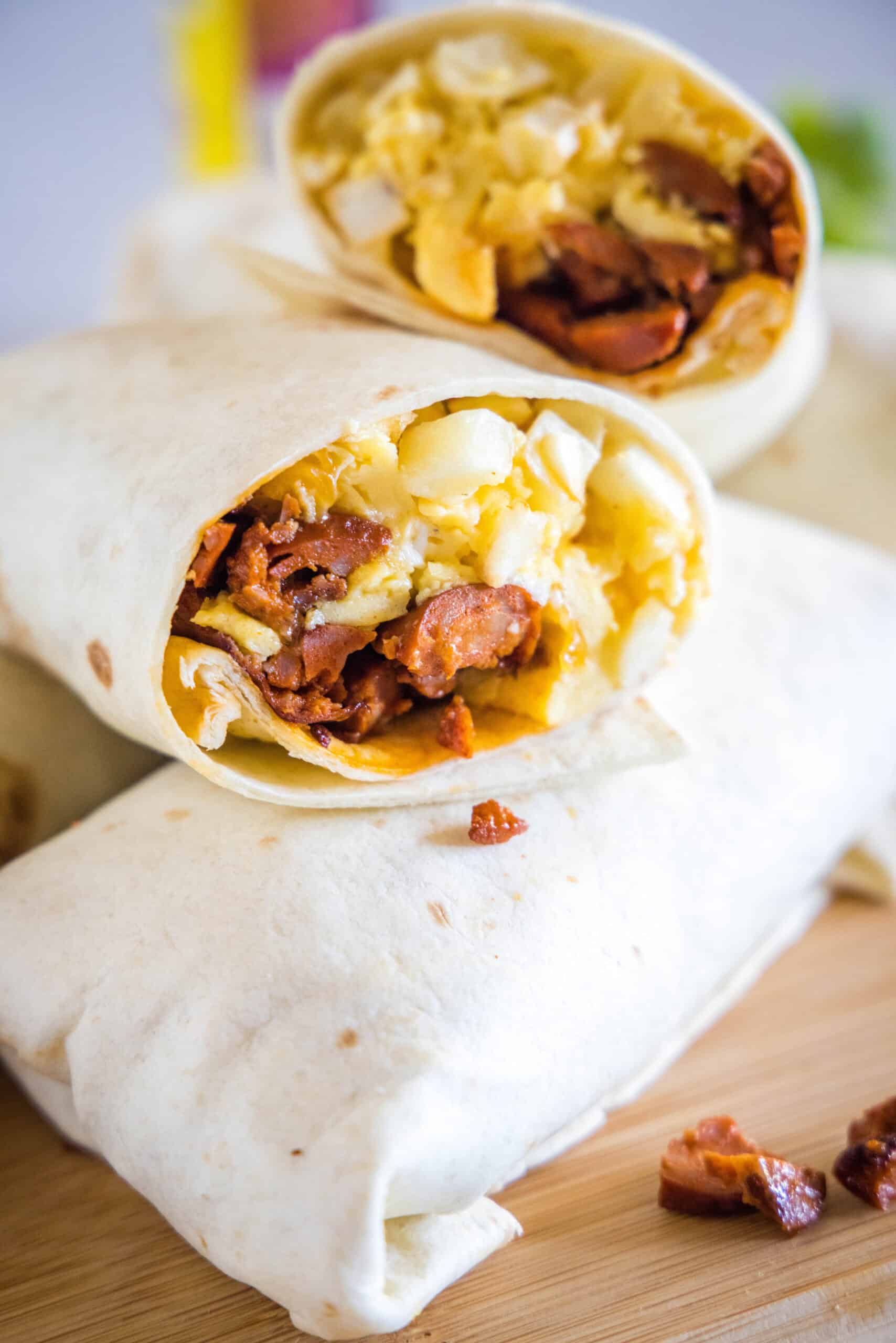 This chorizo breakfast burrito is a cheesy breakfast wrap loaded with crispy hash brown potatoes, spicy chorizo sausage, and fluffy eggs! Two halves of a chorizo breakfast burrito propped up on top of a whole rolled burrito on a wooden cutting board.