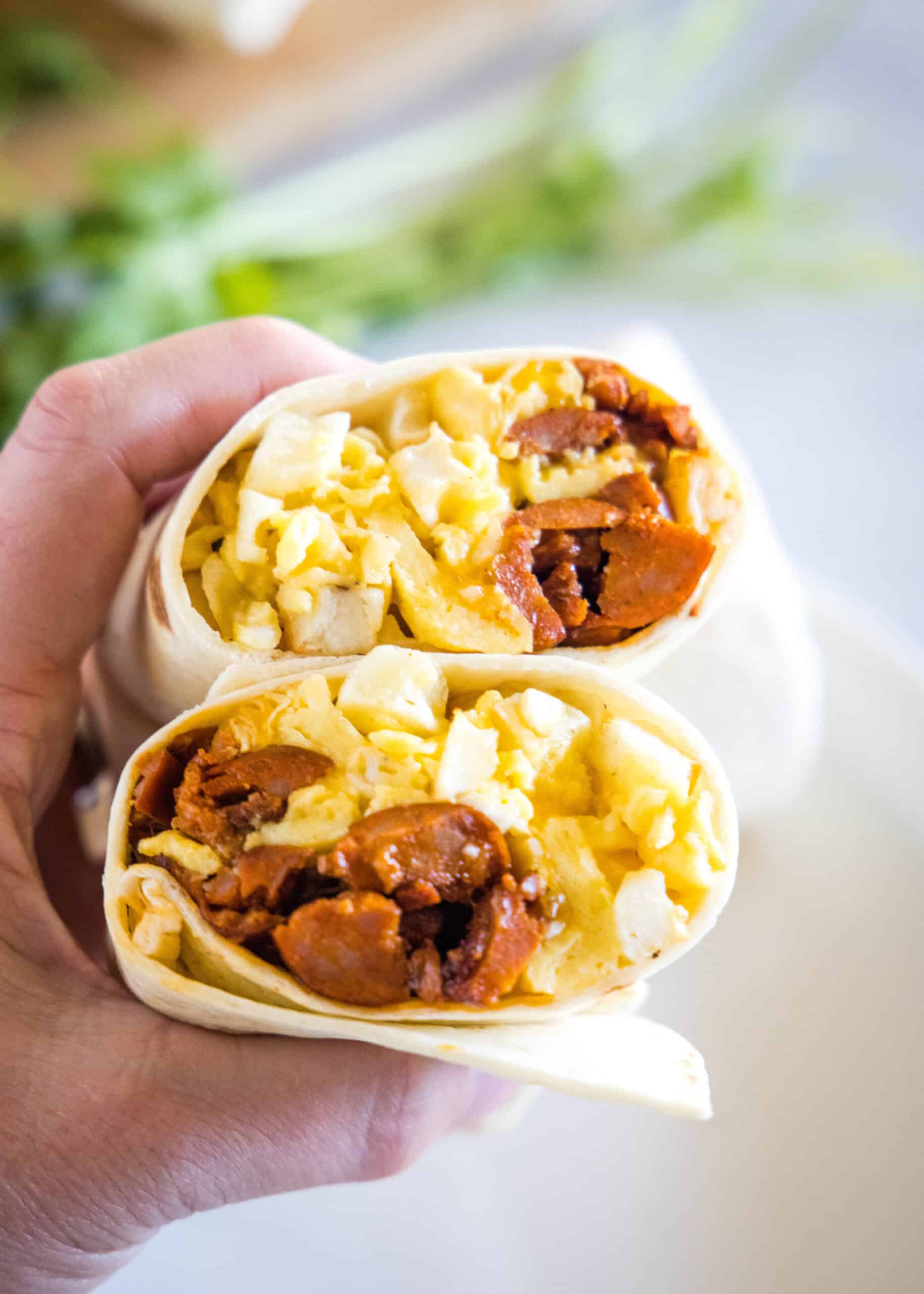 This chorizo breakfast burrito is a cheesy breakfast wrap loaded with crispy hash brown potatoes, spicy chorizo sausage, and fluffy eggs! A hand holding up two halves of a chorizo breakfast burrito, showing the eggs, cheese, potatoes, and chorizo filling inside.