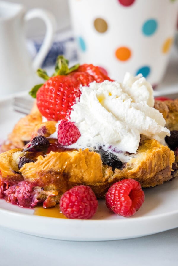 A serving of croissant baked French toast topped with maple syrup, whipped cream, and fresh berries on a plate.