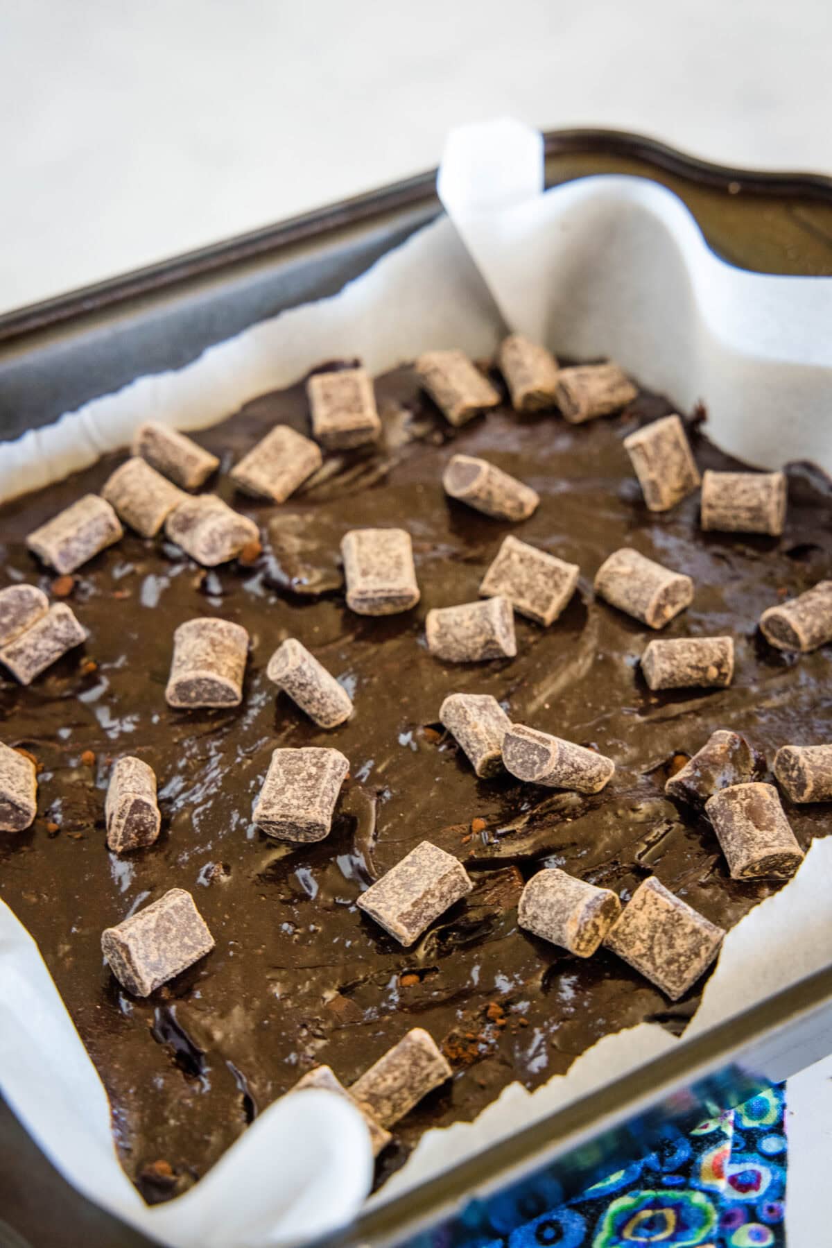 Brownie batter topped with scattered chocolate chunks inside a parchment-lined square pan.