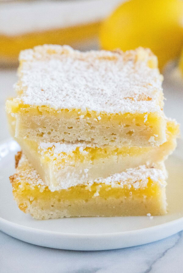 Three square lemon bars stacked on a plate with more bars and lemons behind them