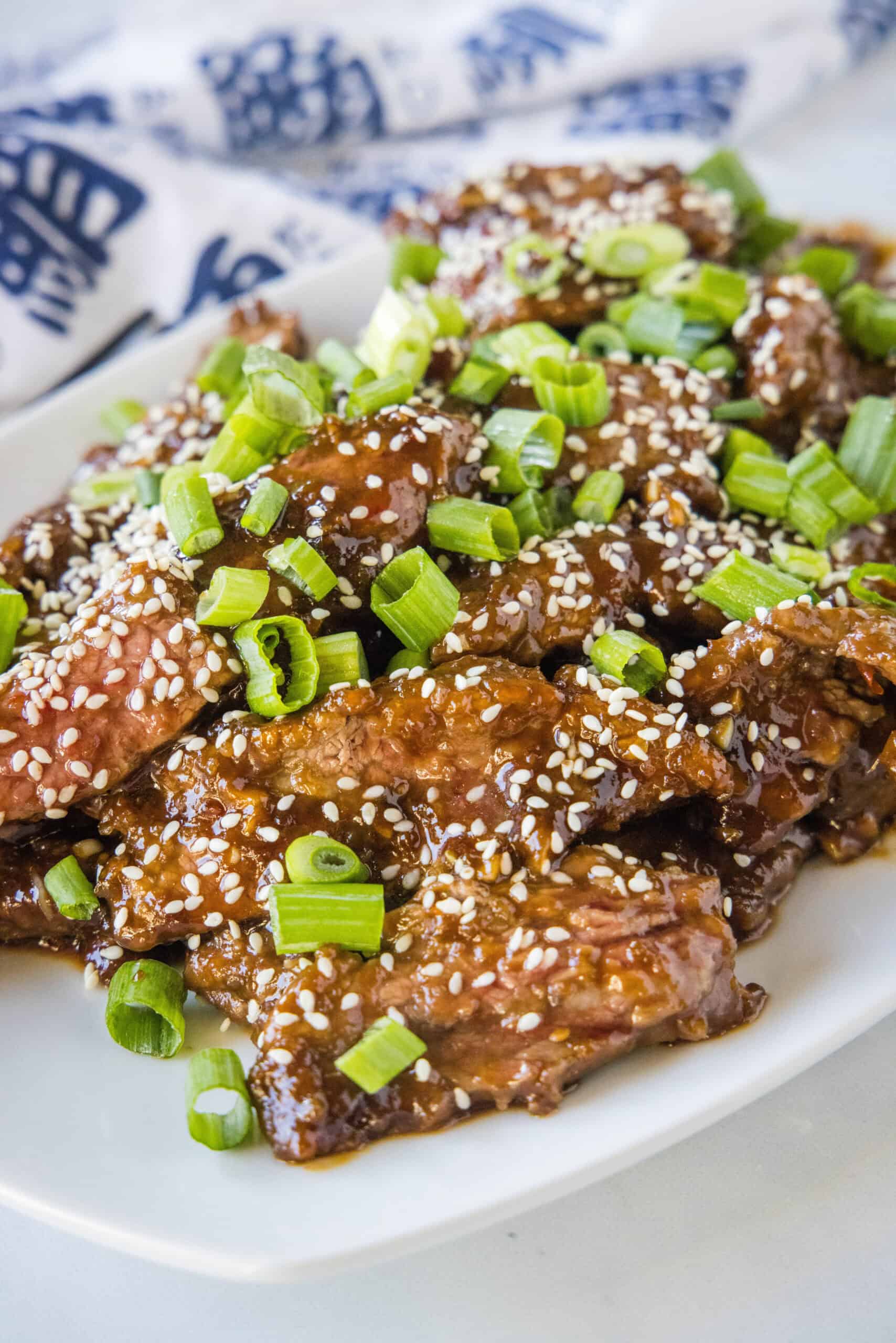 This better-than-takeout Mongolian beef is ready in 25 minutes with crispy, tender steak strips cooked in a sweet and sticky stir-fry sauce. Mongolian beef topped with sesame seeds and green onions on a white platter.