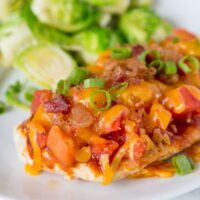 A close up of a plate of food with chicken topped with tomatoes, bacon and melted cheese