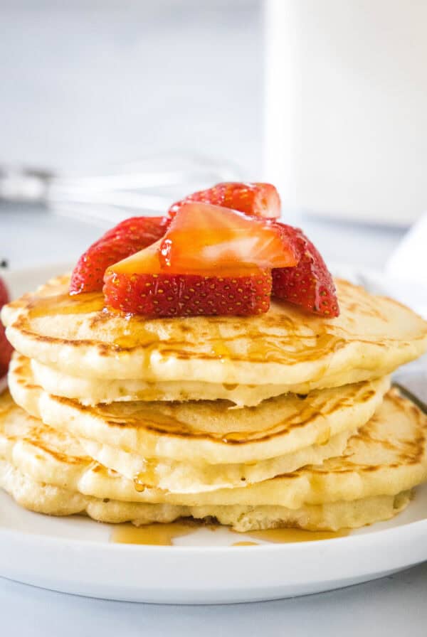 A stack of homemade pancakes topped with fresh strawberries and syrup on a white plate.