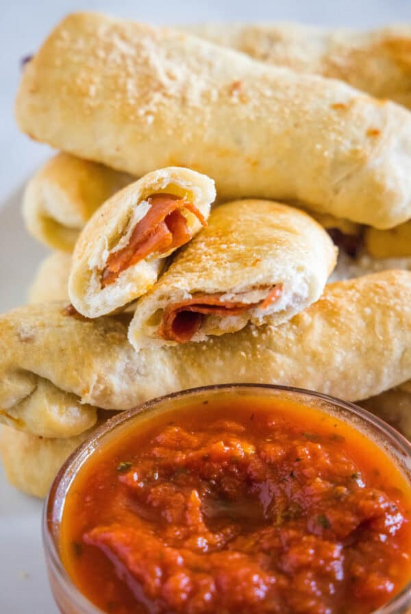 Pepperoni rolls stacked behind a bowl of marinara sauce, with one roll broken in half.