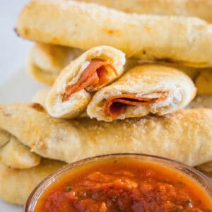 Pepperoni rolls stacked behind a bowl of marinara sauce, with one roll broken in half.