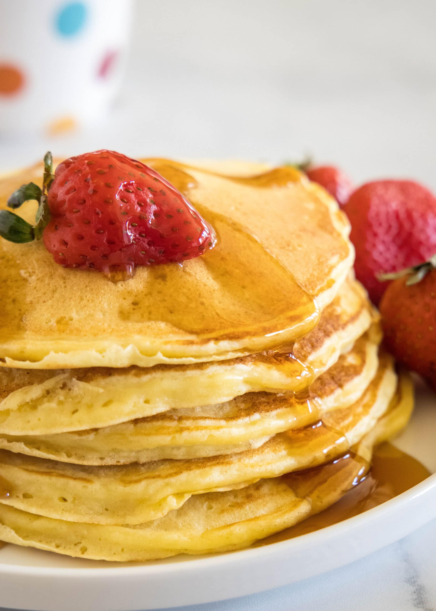 Ricotta pancakes are extra soft, fluffy, and so flavorful! Top these with syrup and a squeeze of lemon for the ultimate breakfast treat! A stack of ricotta pancakes topped with a strawberry and maple syrup on a plate, next to more fresh strawberries.