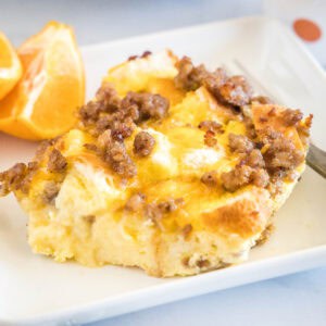 A serving of sourdough breakfast casserole next to orange slices and a fork on a white square plate.