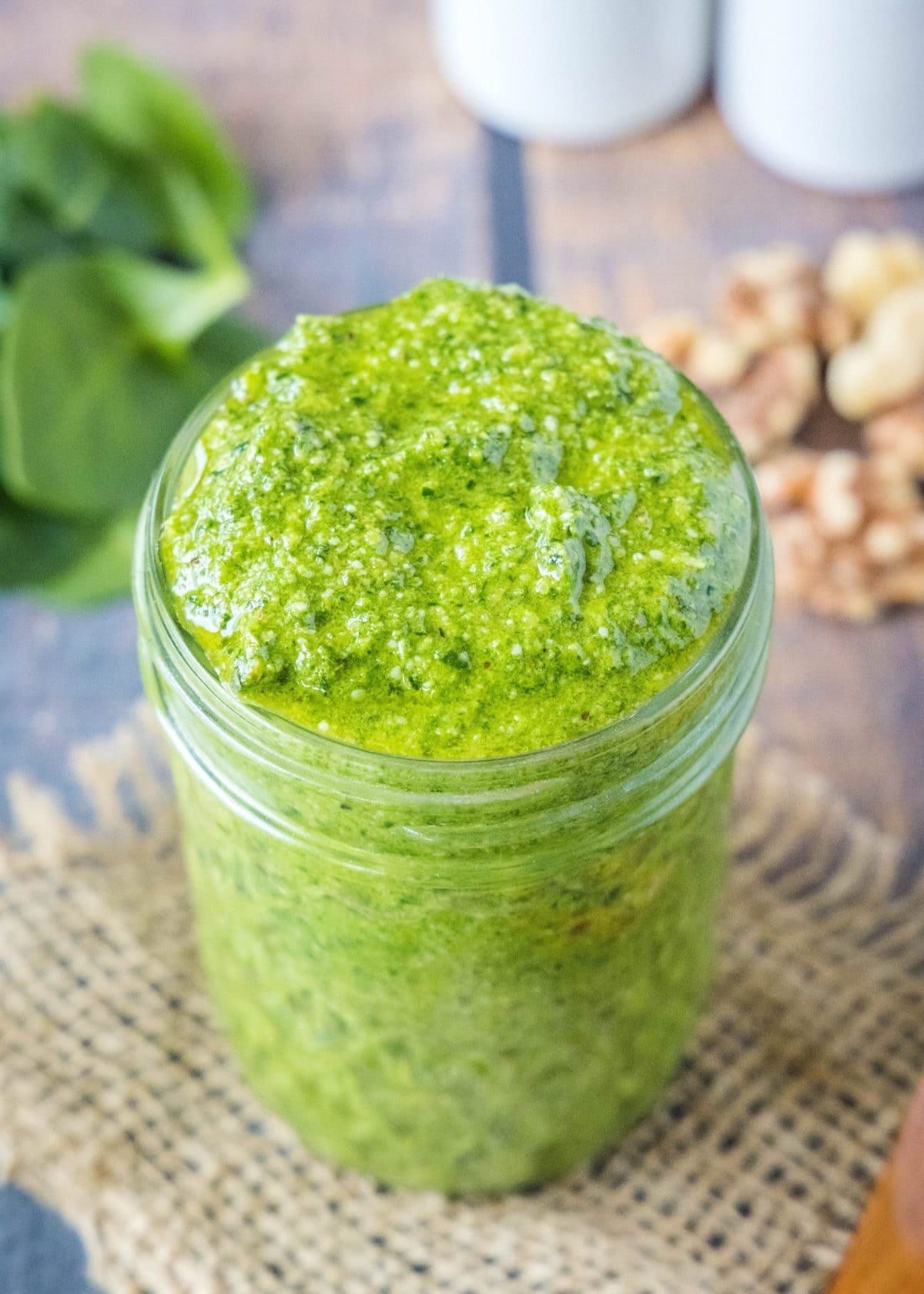 This spinach pesto recipe is healthier and more affordable than a traditional pesto, and it only takes 10 minutes to make! Close up of a jar of pesto with spinach and walnuts in the background