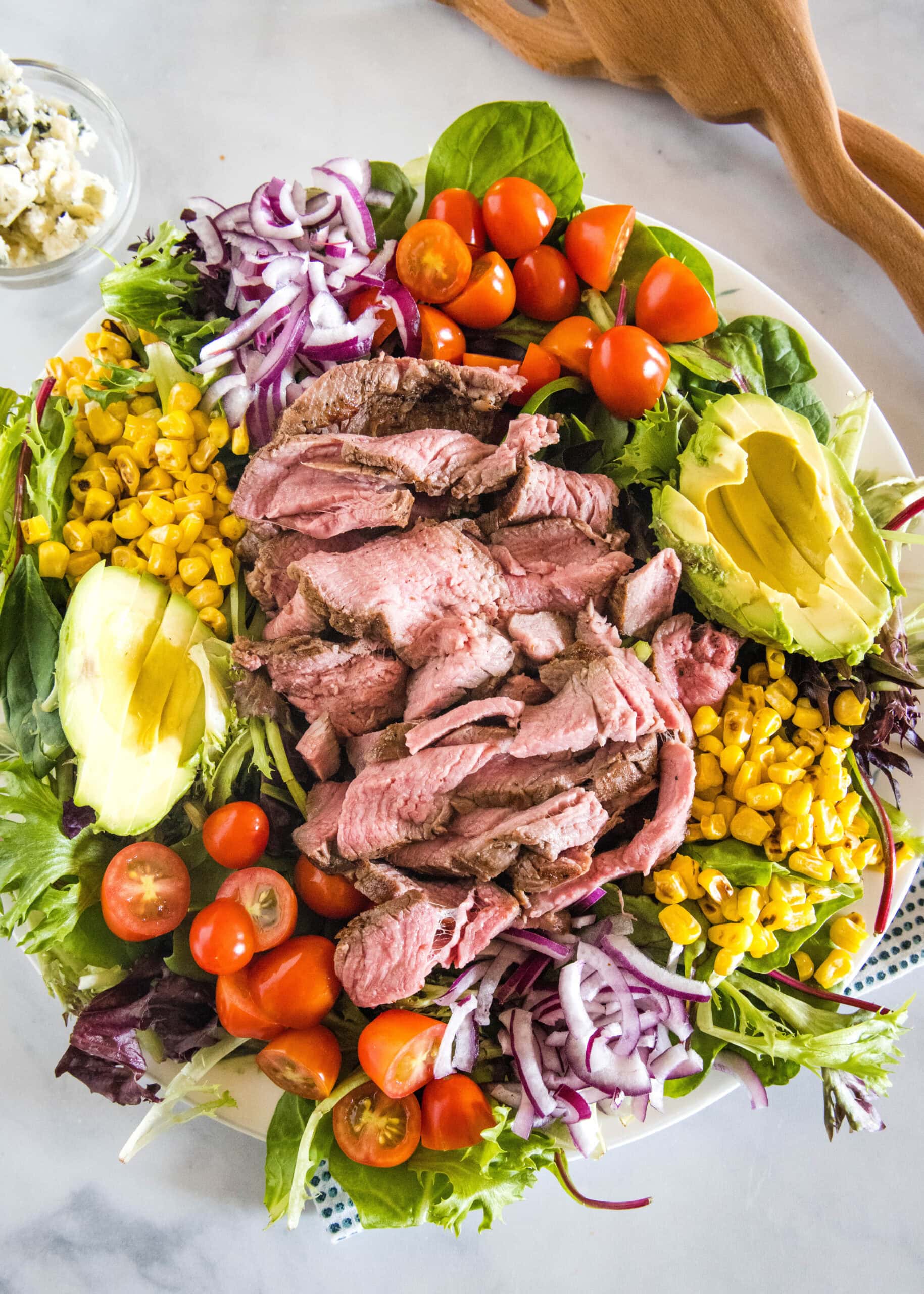 This easy steak salad recipe with fresh summer veggies, avocado, blue cheese, and creamy balsamic dressing is perfect for grilling season! Overhead view of a steak salad on a large platter.