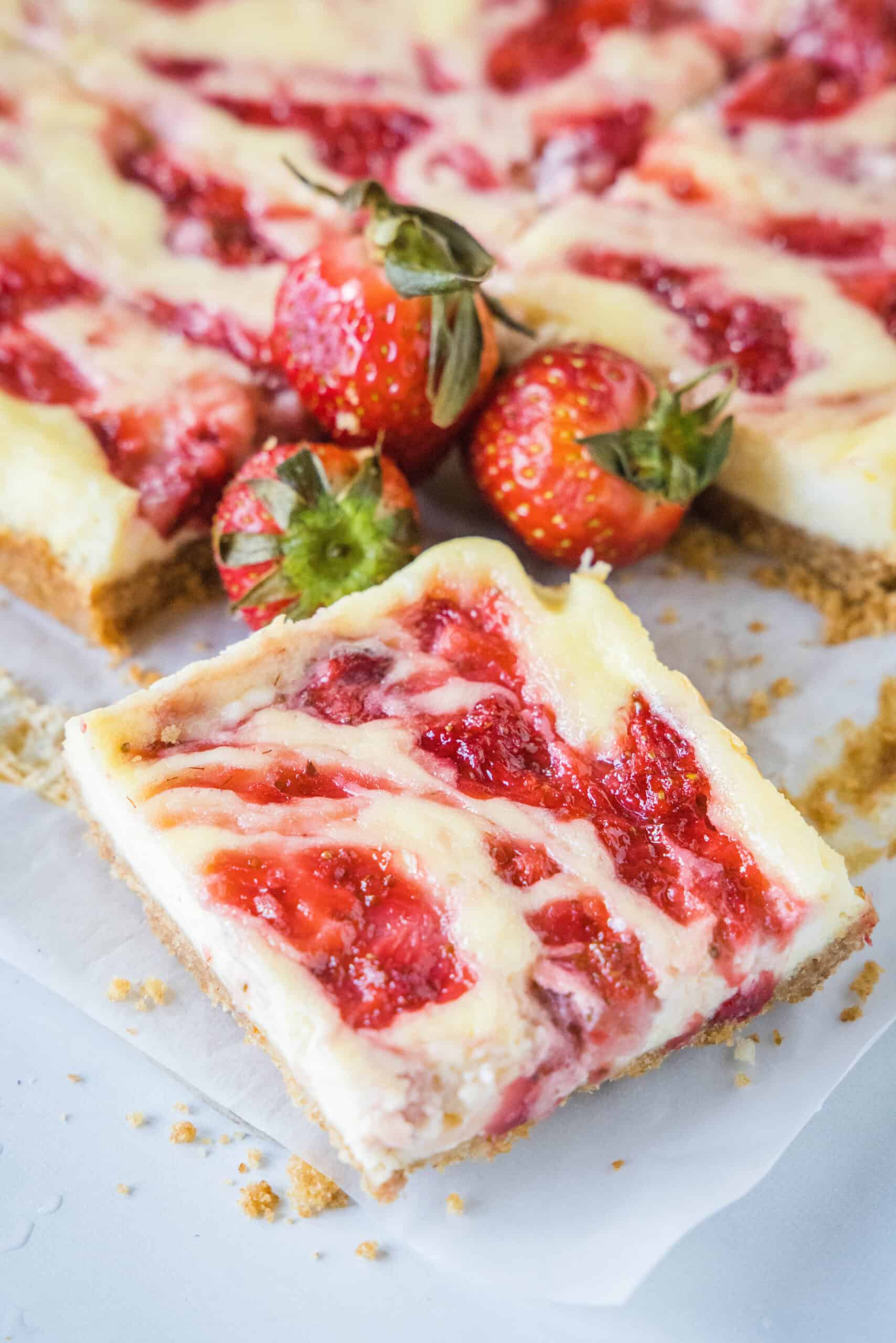 These strawberry cheesecake bars have a tangy cream cheese filling swirled with gooey strawberries, baked on top of a graham cracker crust. Close up of a strawberry cheesecake bar with fresh strawberries and more bars in the background.