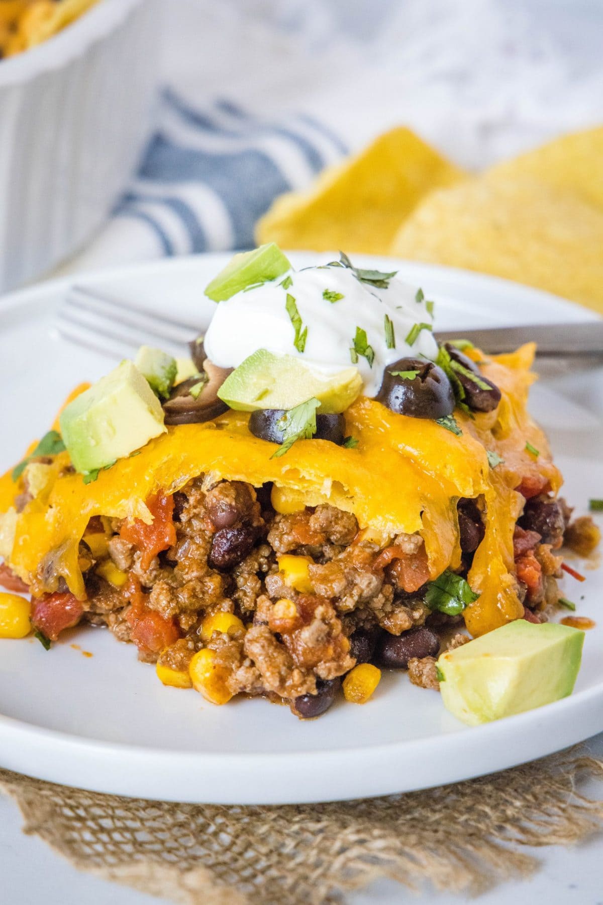 This easy 30-minute taco casserole is loaded with juicy and tender beef, gooey melted cheese, and your favorite taco toppings. A plate with a serving of taco casserole, topped with sour cream, cilantro, olives, and avocado, with chips in the background