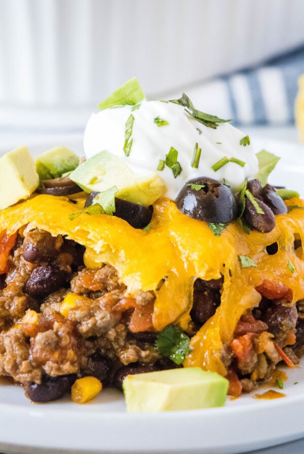 A serving of taco casserole on a plate, topped with sour cream, avocado, cilantro, and olives